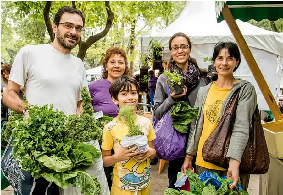 Fotografía de una familia en un mercado al aire libre sosteniendo verduras y plantas; al fondo hay puestos con toldos blancos y árboles.