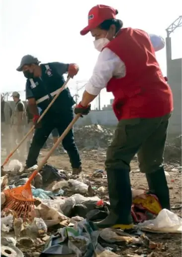 Fotografía de personas con cubre bocas y guantes realizando una campaña de limpieza, recogiendo basura con escobas en un terreno contaminado.