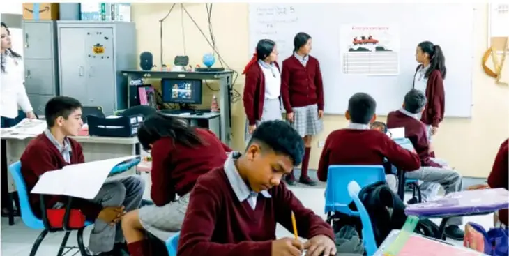 Fotografía de estudiantes de telesecundaria en el aula presentando sus proyectos, con un pizarrón, equipo de cómputo y compañeros escuchando.