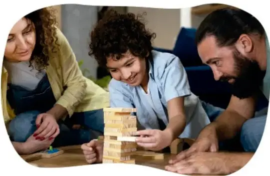 Fotografía de una familia (mujer, niño y hombre barbudo) jugando en el suelo de la sala a torre de bloques de madera estilo Jenga, concentrados y sonrientes.