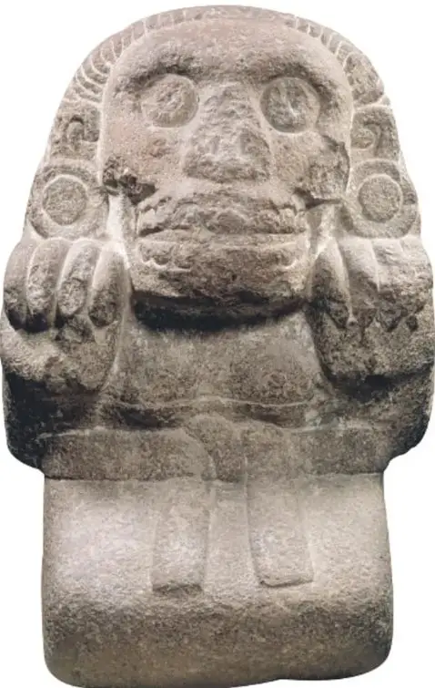 Escultura de piedra de Cihuacóatl: figura con cabeza de calavera o felino con grandes ojos circulares y boca con colmillos, manos que se apoyan frente al pecho y cuerpo rígido de pie.