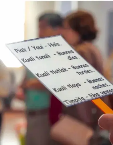 Fotografía de una persona sosteniendo una tarjeta de bienvenida bilingüe con saludos (Piiali/Yeui – Hola; Kuali tonloli – Buenos días; Kuali tiofiok – Buenas tardes; Kuali tiioyo – Buenas noches; Tiimoito – Nos vemos), con público desenfocado al fondo.