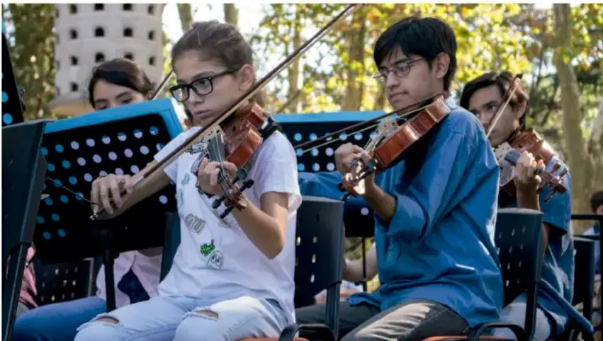 Fotografía de un grupo de estudiantes de secundaria tocando violines al aire libre, sentados frente a atriles de música, en lo que parece una presentación o ensayo de una orquesta escolar. Al fondo se aprecian árboles y una torre (posiblemente la Torre Inclinada de Pisa, como referencia visual).