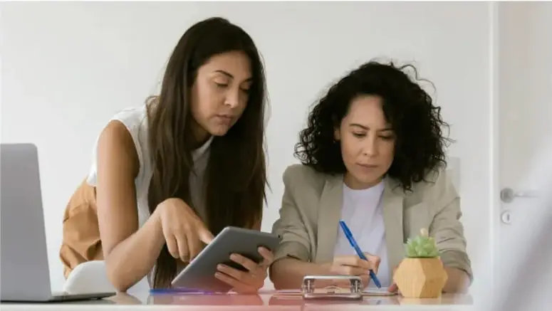 Dos mujeres adultas revisando una tableta y un cuaderno, colaborando en una mesa de trabajo.