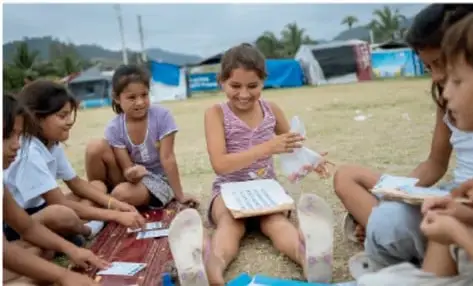 Fotografía de un grupo de niñas y niños sentados en el suelo, al aire libre, leyendo y compartiendo libros. Parecen estar en un entorno comunitario o rural.