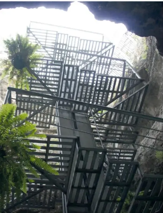 Escaleras metálicas de descenso al interior del géiser, entre rocas y plantas verdes.