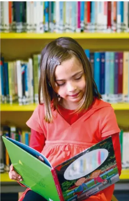 Fotografía de una niña de cabello castaño con una blusa roja, leyendo un libro abierto frente a un estante lleno de libros de colores.