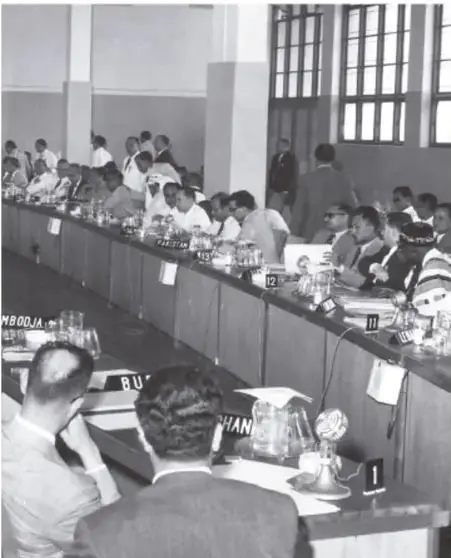 Fotografía en blanco y negro de una sesión de la Conferencia de Bandung, con delegados sentados en una mesa larga y carteles con nombres de países como 'MBOJA' y 'RU'.