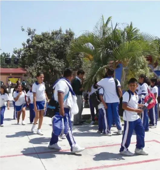 Fotografía de estudiantes de telesecundaria con uniforme blanco y azul caminando en el patio de su escuela.