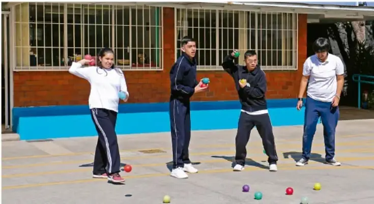 Fotografía de cuatro jóvenes en el patio de una escuela realizando una actividad física con pelotas de colores; tres están en posición de lanzamiento y un adulto los observa.