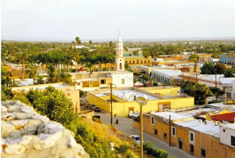 Vista panorámica de El Fuerte, Sinaloa: casas de techos de colores, torre blanca de iglesia, palmeras y un cerro rocoso; tonos cálidos del norte de Sinaloa.