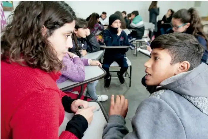 Fotografía de estudiantes de primaria sentados en un salón de clases, conversando y participando en una actividad grupal.