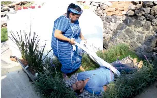 Fotografía de una mujer con vestimenta tradicional azul moliendo maíz en un metate, junto a un hombre recostado sobre un petate; al fondo, un muro de piedra y plantas, escena de la vida cotidiana rural mexicana.