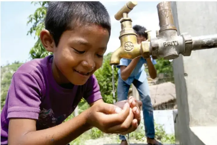 Fotografía de un niño bebiendo agua con las manos junto a una llave de paso, con otro niño en el fondo; ilustra la importancia del acceso al agua.