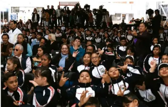 Fotografía de un grupo numeroso de estudiantes con uniforme escolar y personas adultas reunidos en un espacio institucional, muchos levantando la mano en señal de participación.