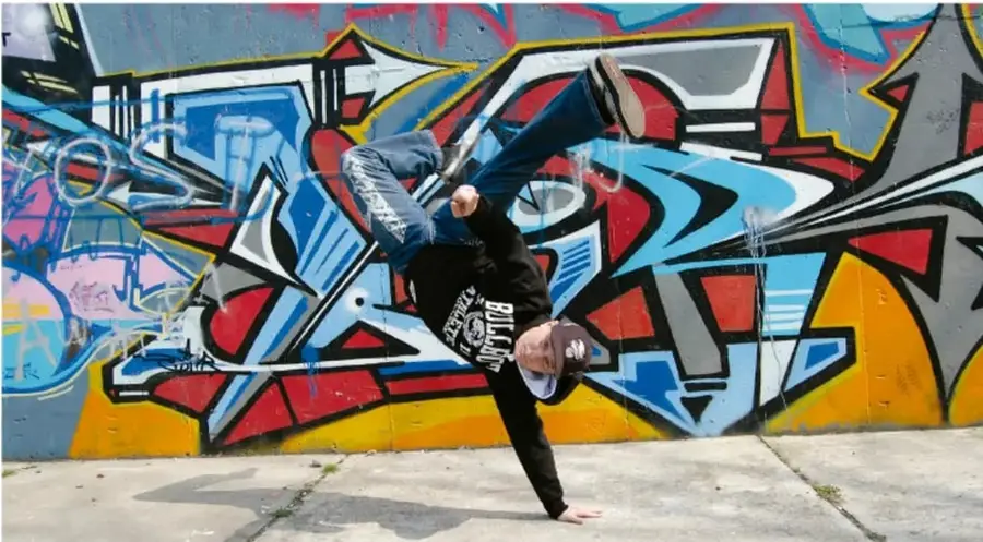 Fotografía de un bailarín de break dance ejecutando una figura de una mano frente a un muro cubierto de grafiti colorido (azul, rojo, amarillo, negro).