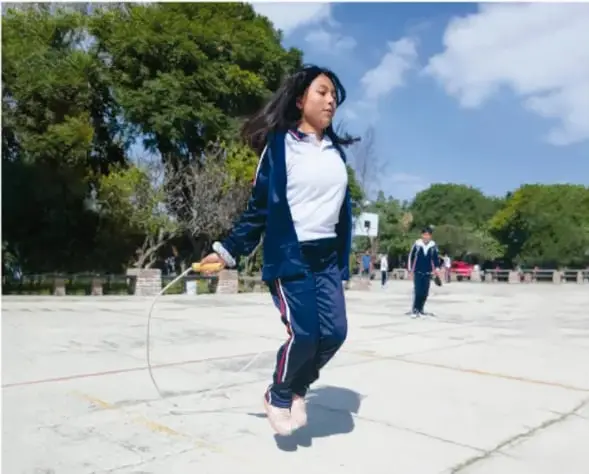 Fotografía de una estudiante en uniforme saltando la cuerda en el patio de una escuela, con árboles y otros estudiantes al fondo, ilustrando la actividad física en el entorno escolar.