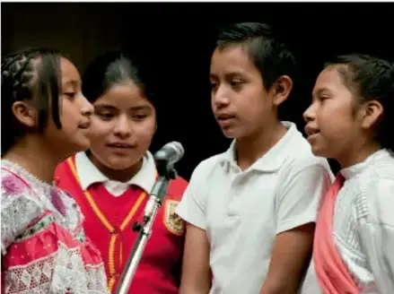 Fotografía de cuatro niñas y niños indígenas ante un micrófono, aparentemente participando en un acto o presentación cultural.