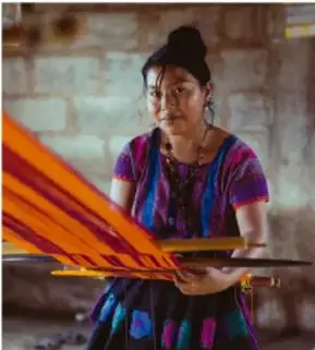Mujer indígena con vestimenta tradicional sostiene un telar de cintura con hilos naranjas.
