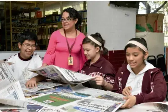 Fotografía de un grupo de estudiantes junto con una docente observando y comentando periódicos impresos en un espacio escolar.