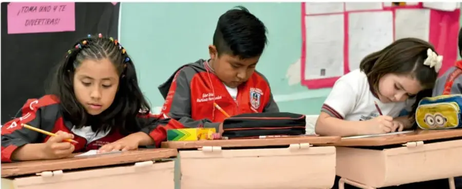 Fotografía de tres estudiantes de primaria trabajando en sus pupitres dentro del aula; una niña escribe, un niño revisa un estuche y otra niña dibuja o escribe. En la pared hay un cartel rosado con la frase '¡TOMA UNO Y TE DIVERTIRÁS!'.