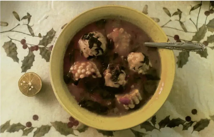 Fotografía cenital de un plato tradicional servido en un tazón amarillo con caldo, acompañado de una rodaja de cítrico, sobre un mantel con motivos de hojas y bayas.