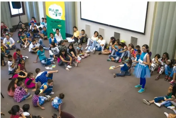Fotografía de una asamblea escolar: estudiantes de primaria sentados en círculo en el suelo de un salón amplio, con una niña de pie al frente dirigiendo la actividad; al fondo hay una proyección y un cartel verde con la leyenda 'Diálogos en la escuela'.