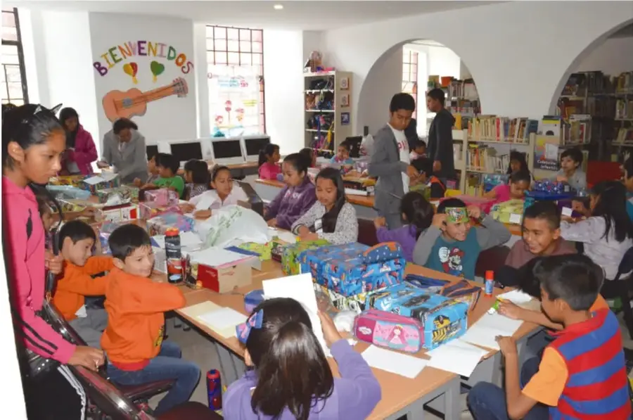 Fotografía de un aula o biblioteca escolar con niñas y niños trabajando en mesas con cajas, papel y materiales; al fondo hay estantes con libros y un letrero que dice «BIENVENIDOS».