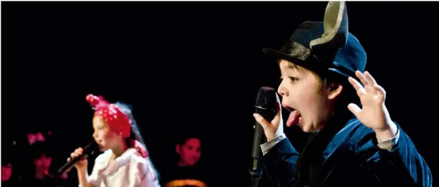 Fotografía de dos niños sobre un escenario oscuro: una niña con peluca roja canta frente a un micrófono y un niño con sombrero y saco azul canta con gesto expresivo; al fondo se distingue un adulto.