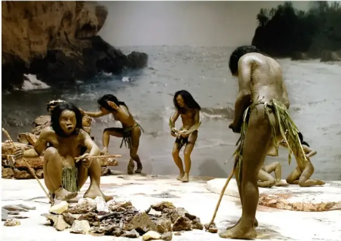 Fotografía o recreación de seres humanos antiguos en una zona costera rocosa, trabajando con piedras y madera; ilustra la vida de los antiguos habitantes.