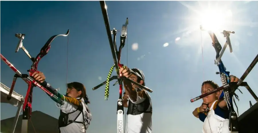 Fotografía de tres estudiantes de secundaria practicando tiro con arco al aire libre, con el sol a contraluz; sostienen arcos deportivos y muestran concentración y postura de puntería.