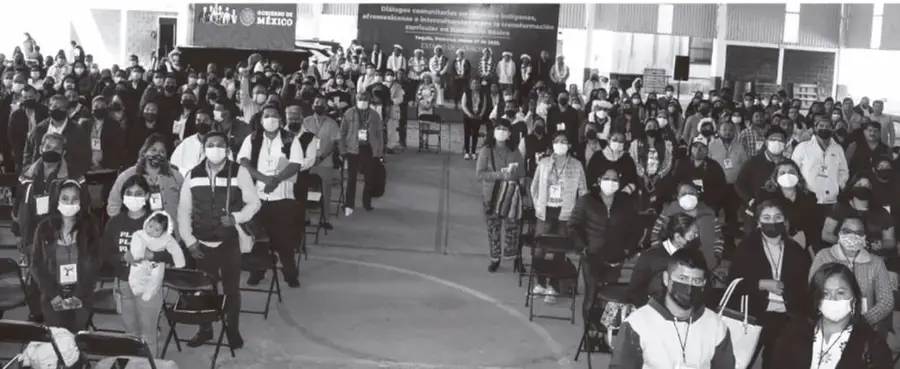 Fotografía en blanco y negro de una asamblea o consulta comunitaria en un patio escolar: multitud de personas sentadas y de pie, muchas con cubrebocas y credenciales, con un escenario al fondo que muestra el logotipo 'Gobierno de México' y una manta con texto parcialmente legible; ilustra la participación de las familias y la comunidad.