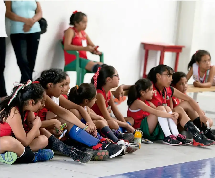 Grupo de estudiantes de secundaria con uniforme deportivo rojo sentados en el piso y en las gradas de una cancha techada, atentos a una actividad; una persona adulta está de pie al fondo.