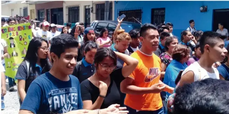 Fotografía de una marcha o manifestación juvenil en la calle. Se observa a un grupo numeroso de adolescentes y jóvenes caminando, algunos con pancartas y carteles. Al frente destaca un joven con playera azul con la palabra 'YANKEE' y otro con playera naranja. El ambiente es de participación y movilización pacífica.