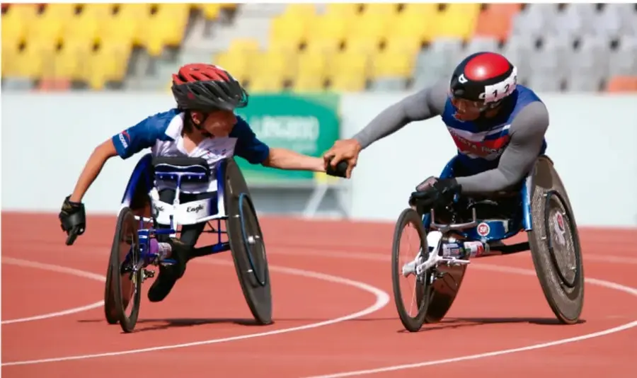 Fotografía de dos atletas en silla de ruedas de competición dándose las manos en una pista de atletismo roja, con gradas amarillas de fondo; representan la inclusión y la camaradería deportiva.