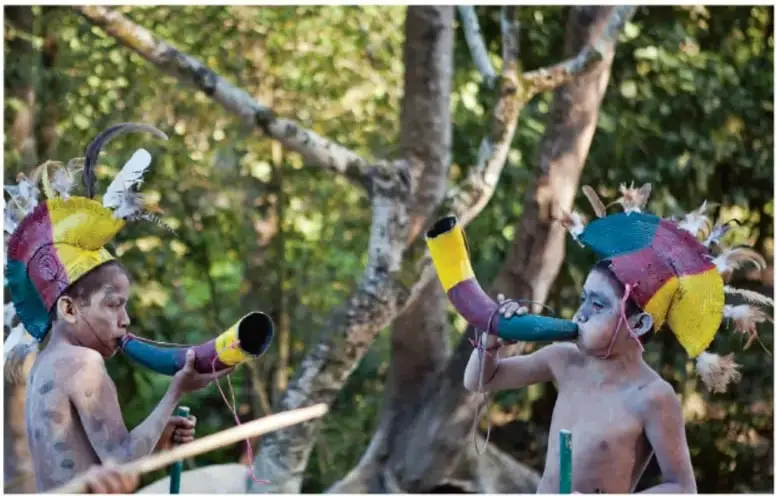 Fotografía de dos niños con penachos de plumas y pintura corporal, sosteniendo tubos pintados de colores, en un entorno arbolado; corresponde a una escena del Festival de Danza de los Mecos en Hidalgo.