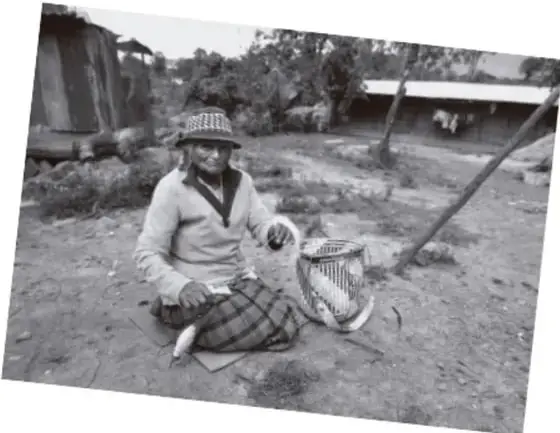 Fotografía en blanco y negro de una mujer indígena sentada en el suelo tejiendo un canasto, con vestimenta tradicional y paisaje rural de fondo.