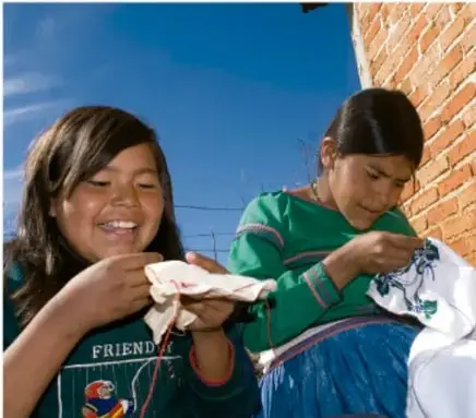 Fotografía de dos niñas/adolescentes indígenas bordando o trabajando textiles, sonriendo, frente a una pared de ladrillo.