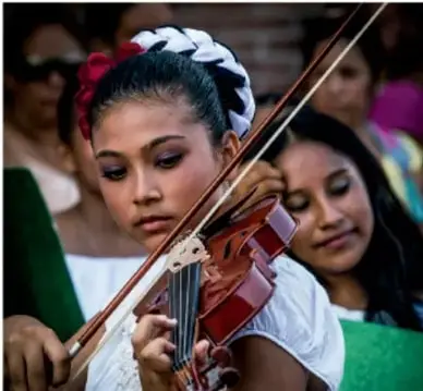 Fotografía de una joven tocando el violín, con flores en el cabello, rodeada de otras personas; ilustra las manifestaciones artísticas y la transmisión cultural.