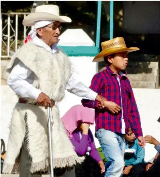 Fotografía de un anciano con sombrero y bastón junto a un niño con sombrero de paja, posiblemente transmitiendo tradiciones orales en un contexto comunitario.