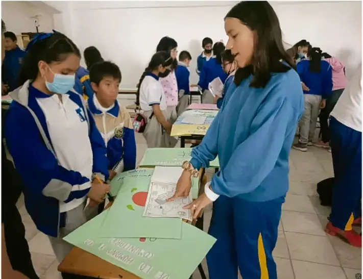 Fotografía de estudiantes de telesecundaria en el aula trabajando con cartulinas y materiales en mesas; una estudiante mayor instruye a dos compañeros; crédito: Luis Eduardo García Pobal, TOMÁTIX.