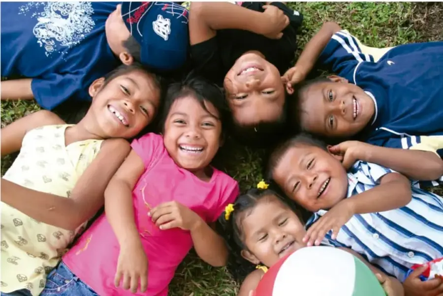 Fotografía de un grupo de niños y niñas acostados en el pasto, sonriendo y formando un círculo con sus cabezas. Algunos usan playeras de colores y uno sostiene un balón. Transmiten alegría y trabajo en equipo.
