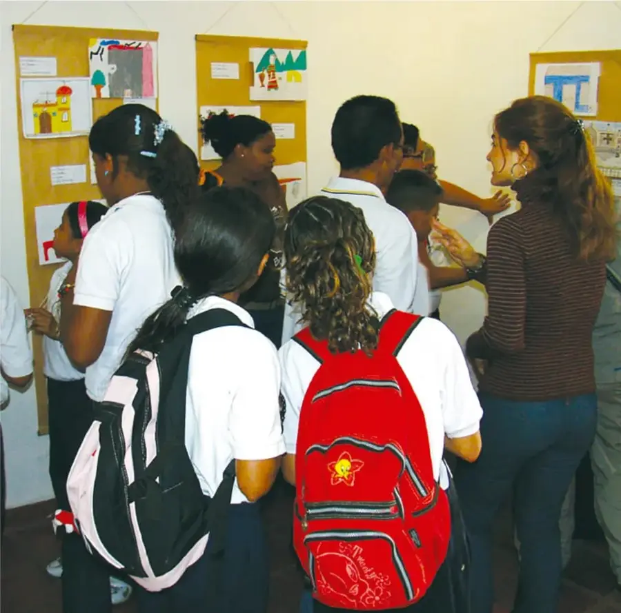 Estudiantes de primaria con uniforme blanco y mochilas observan una galería de trabajos escolares colocados en paneles de madera; una maestra de suéter rayado dialoga con ellos.