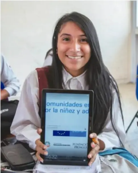 Fotografía de una adolescente sonriente con cabello largo, vestida con camisa blanca y suéter, que sostiene una tablet encendida; en la pantalla se distingue texto relacionado con comunidades de la niñez y adolescencia; parece estar en un aula.