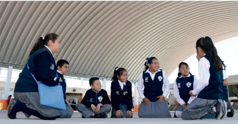 Fotografía de un grupo de estudiantes de primaria sentados en el suelo bajo una estructura metálica, conversando o ensayando.