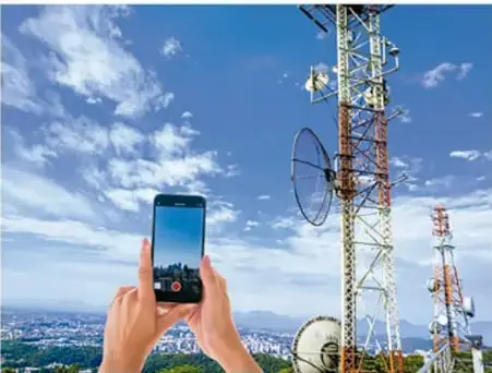 Fotografía de una persona sosteniendo un teléfono móvil frente a una gran torre de antenas de telecomunicaciones bajo un cielo nublado.