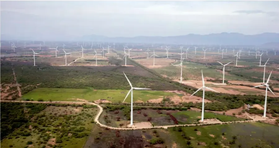 Fotografía aérea de un parque eólico con numerosos aerogeneradores distribuidos en un paisaje árido y semiárido, con vegetación baja, caminos de tierra y montañas al fondo.