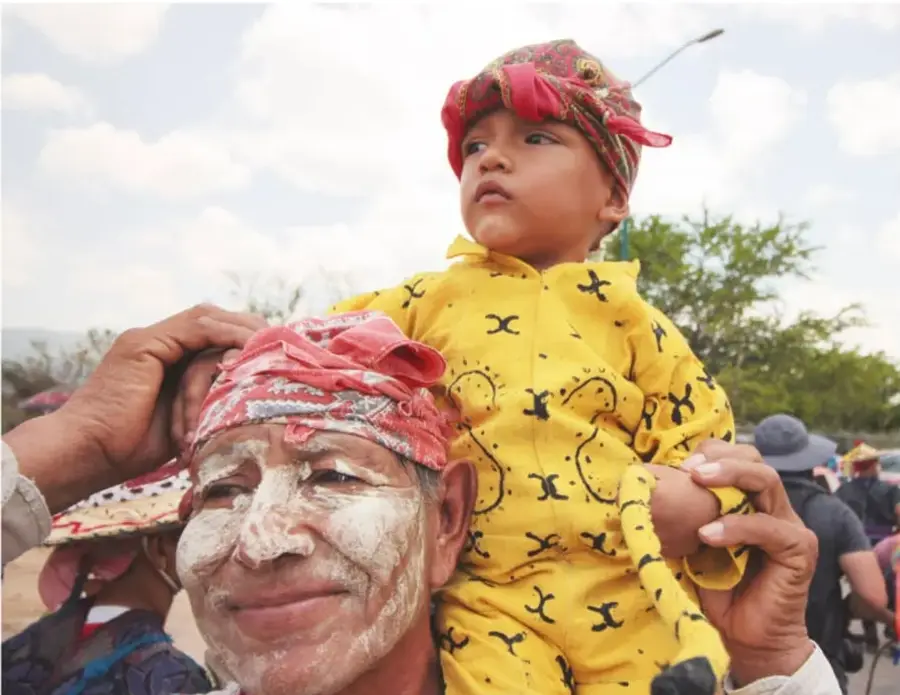 Fotografía a color: un hombre con la cara pintada de blanco y paliacate rojo carga sobre sus hombros a un niño con traje amarillo de manchas negras (parecido a un jaguar o mono) y paliacate rojo; al fondo se ve gente con sombreros y vegetación, en ambiente de fiesta tradicional.