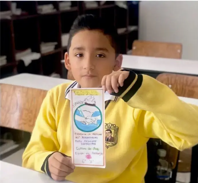 Fotografía de un estudiante con uniforme amarillo sosteniendo un tríptico titulado 'Piénsalo la próxima vez' con dibujos de manos, una paloma y el lema 'Cultura de Paz'.