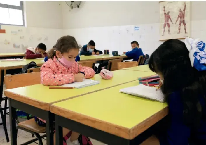 Fotografía de un aula escolar con niñas y niños trabajando en sus pupitres. Una niña con cubrebocas rosa escribe en su cuaderno. Al fondo hay un cartel con figuras humanas.
